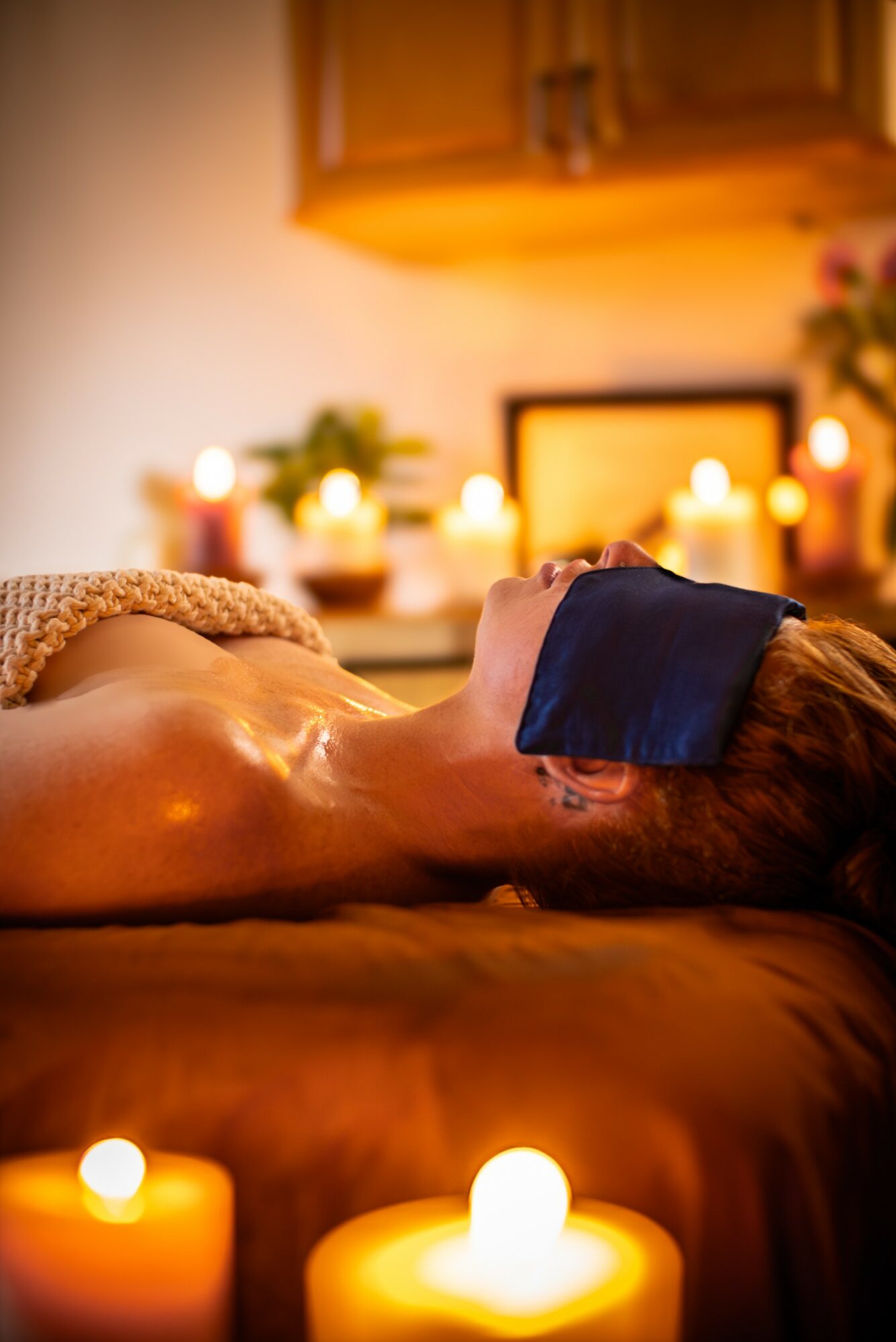 Person lying face up on massage table with eyes covered, surrounded by lit candles and warm lighting in a spa setting.
