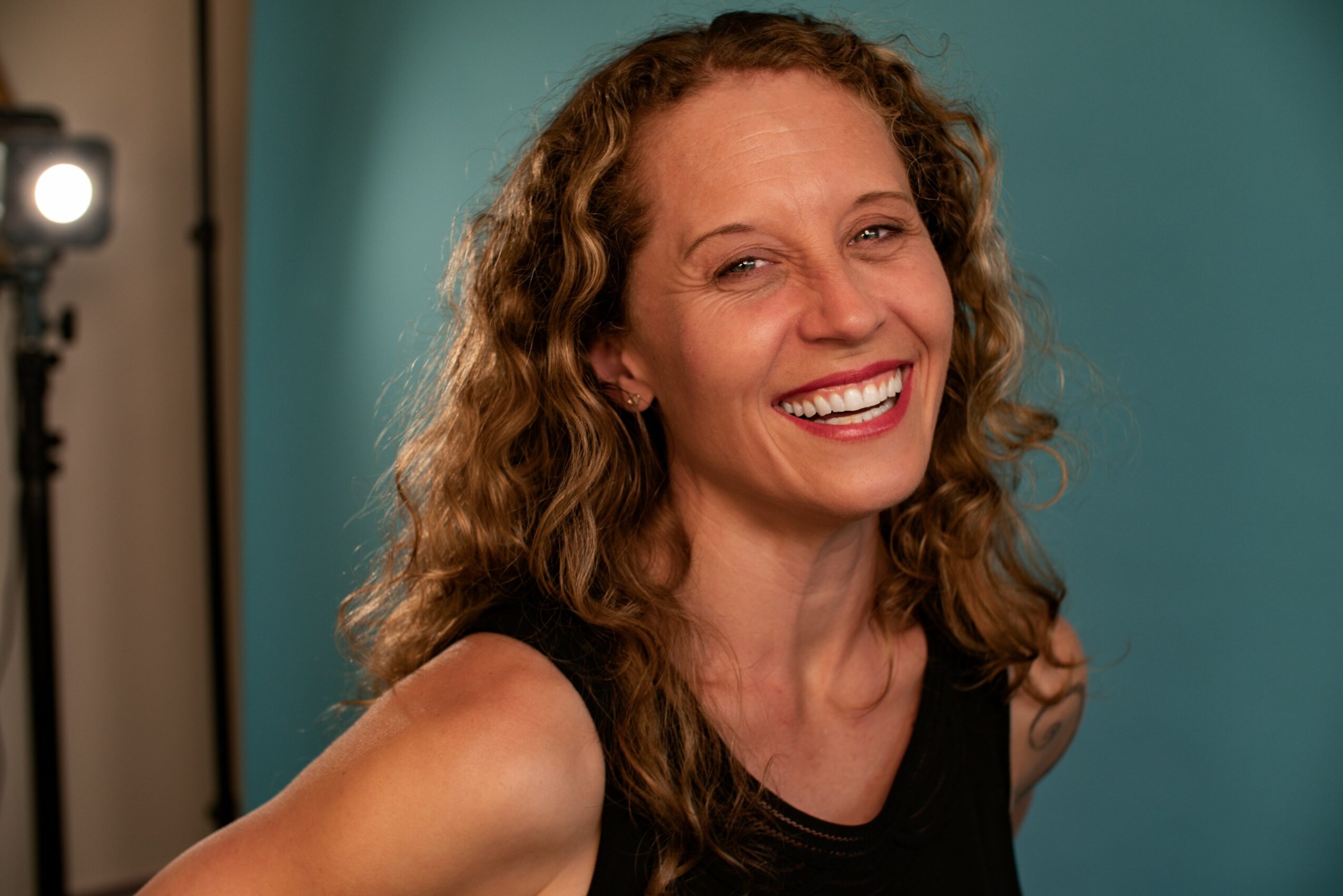 Woman with curly hair smiling, wearing a black top, against a blue background.