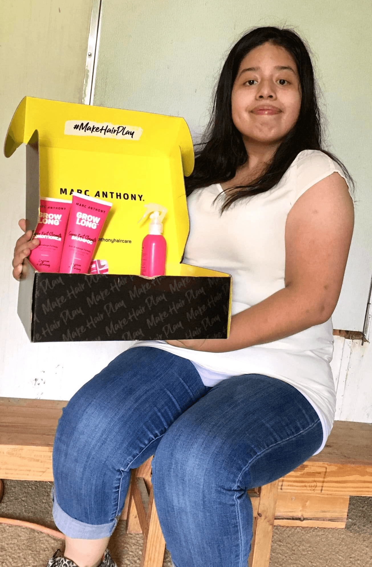 Young girl sitting on a wooden bench holding a yellow and black box with pink bottles inside, smiling at the camera.