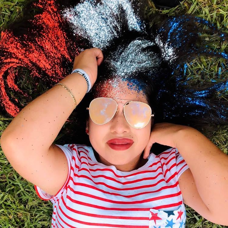 Person lying on grass with colorful hair, wearing sunglasses and a striped shirt, relaxing outdoors.