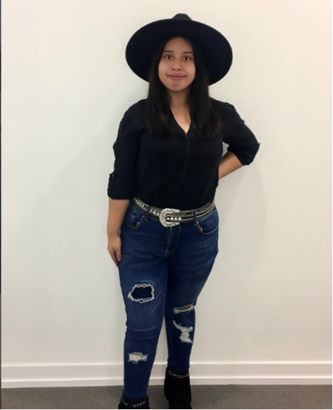 Young woman wearing a large black hat, black top, and ripped jeans, standing against a plain white wall.