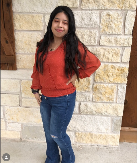 Young woman with long dark hair in a red sweater and blue jeans standing against a stone wall, smiling.