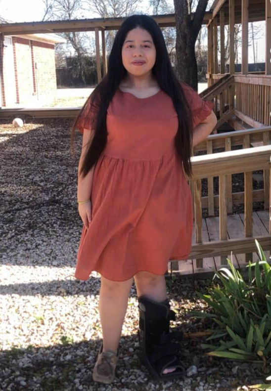 Young woman with long dark hair standing outdoors near a wooden railing and plants, wearing a reddish-brown dress and boots.