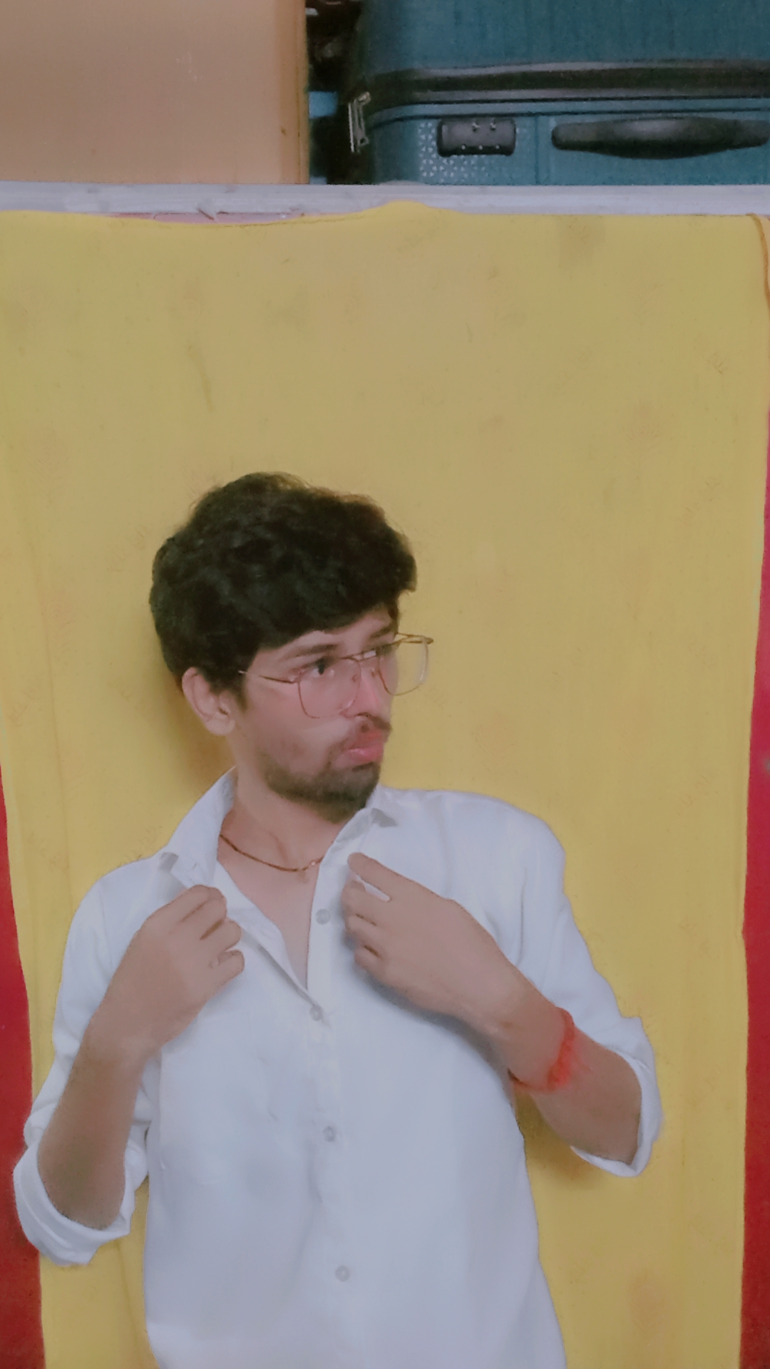 Young man with glasses and curly hair wearing a white shirt, adjusting his collar, standing against a yellow background.
