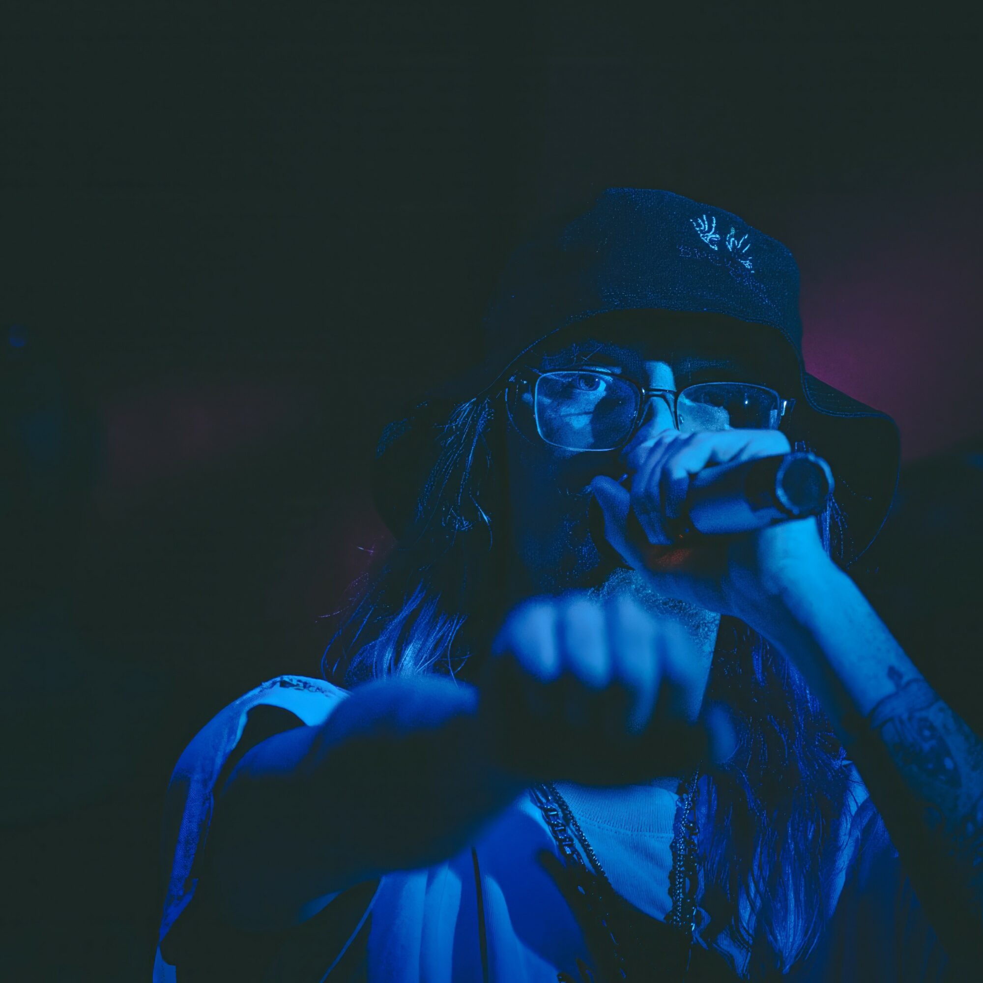 Person wearing glasses and a hat, holding a microphone, in a dark setting with blue lighting.