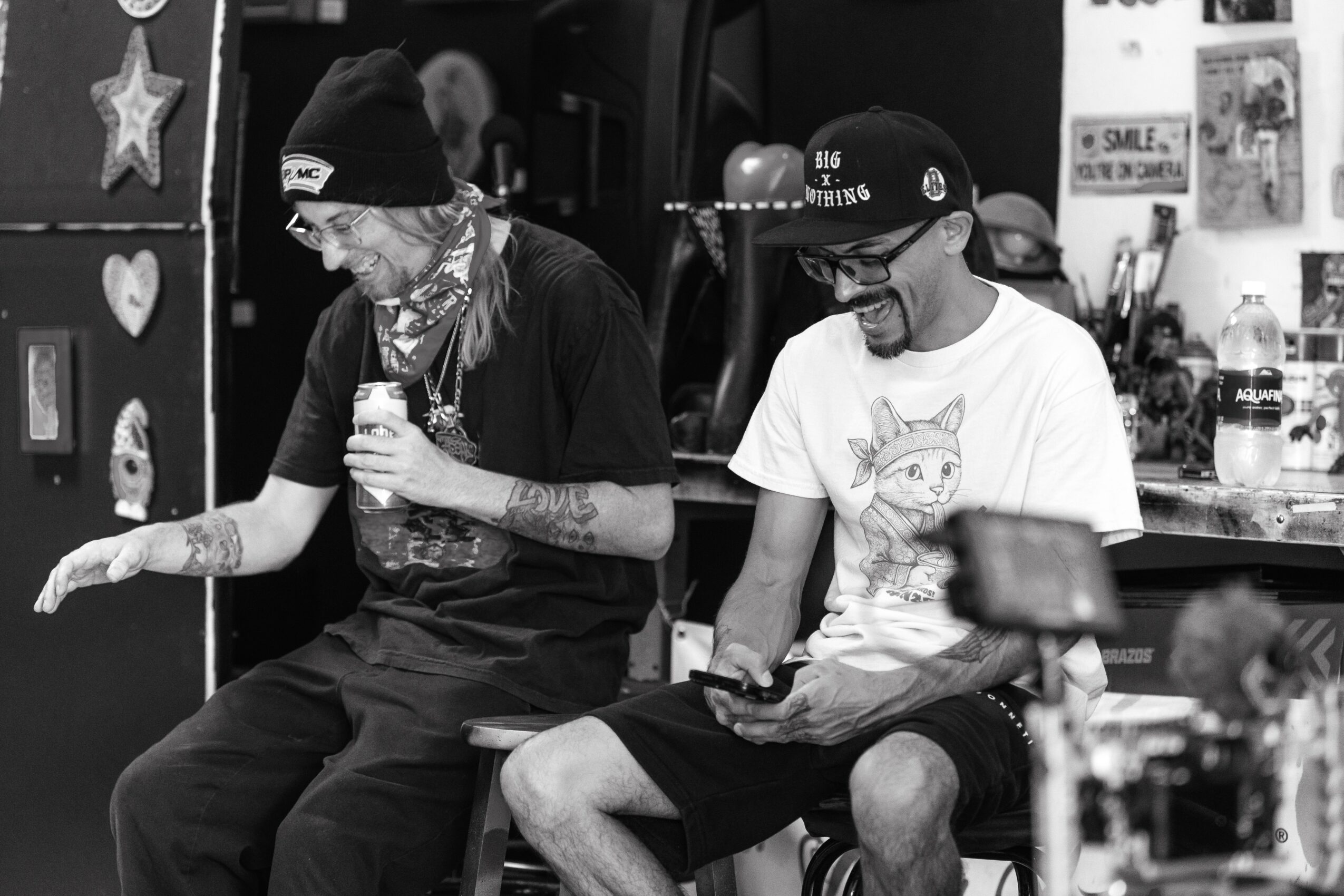 Two people sitting on a bench, one holding a drink and the other looking at a phone, in a room with various items.