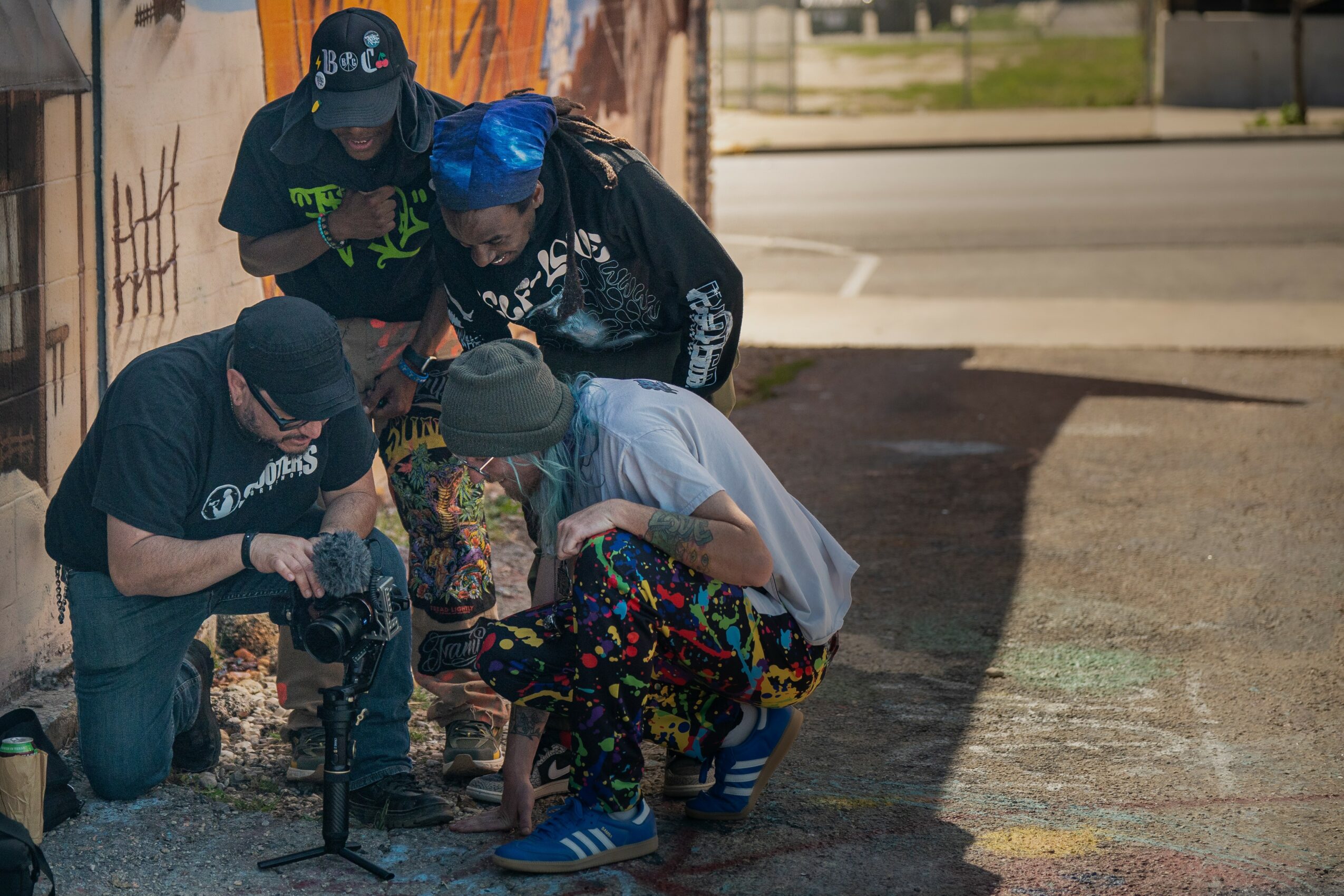 Four people gathered around a camera on a sidewalk, with a wall and street in the background.