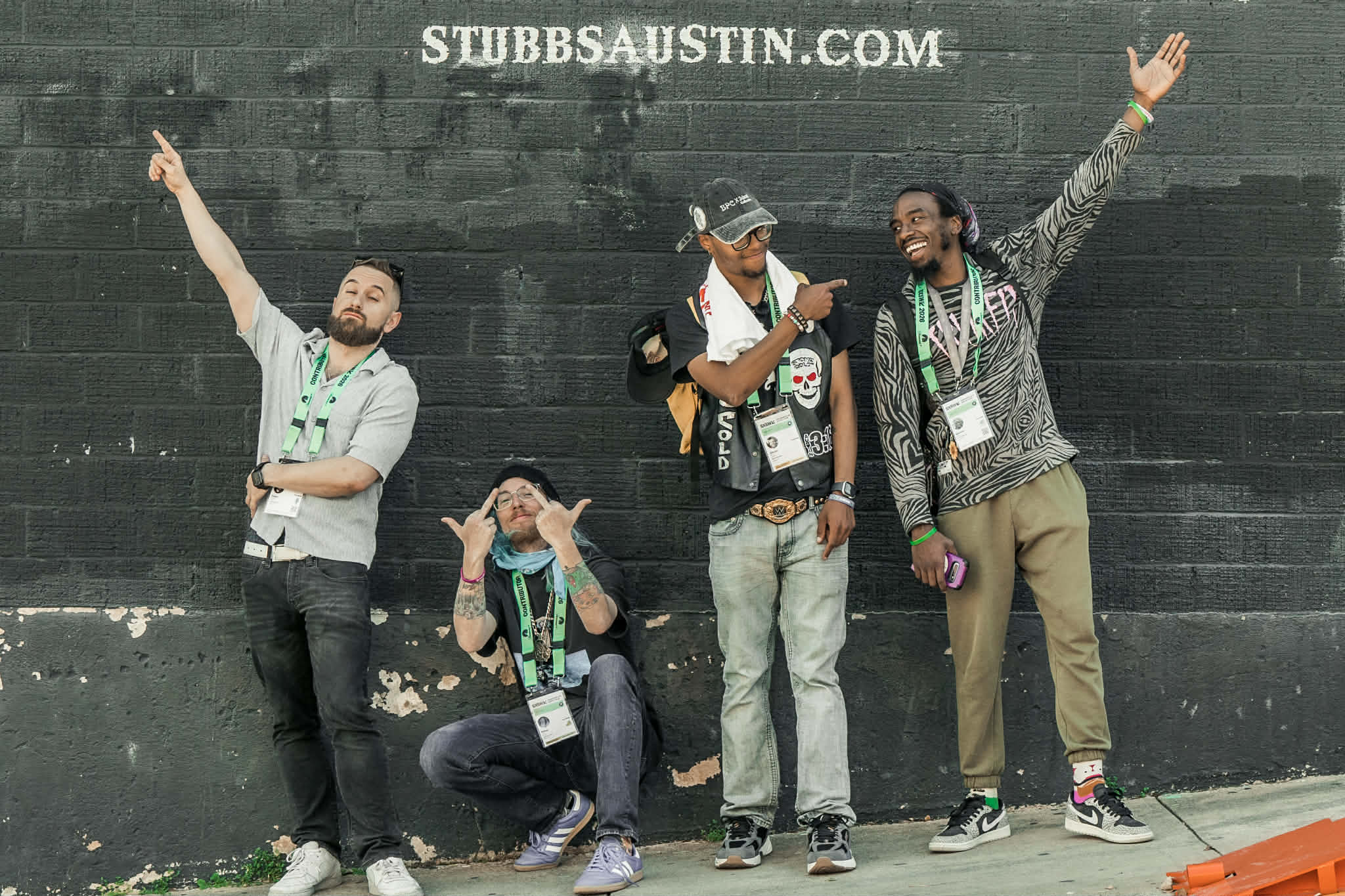 Four people standing and sitting against a black wall, some raising hands, one person covering ears, casual clothing, outdoors.