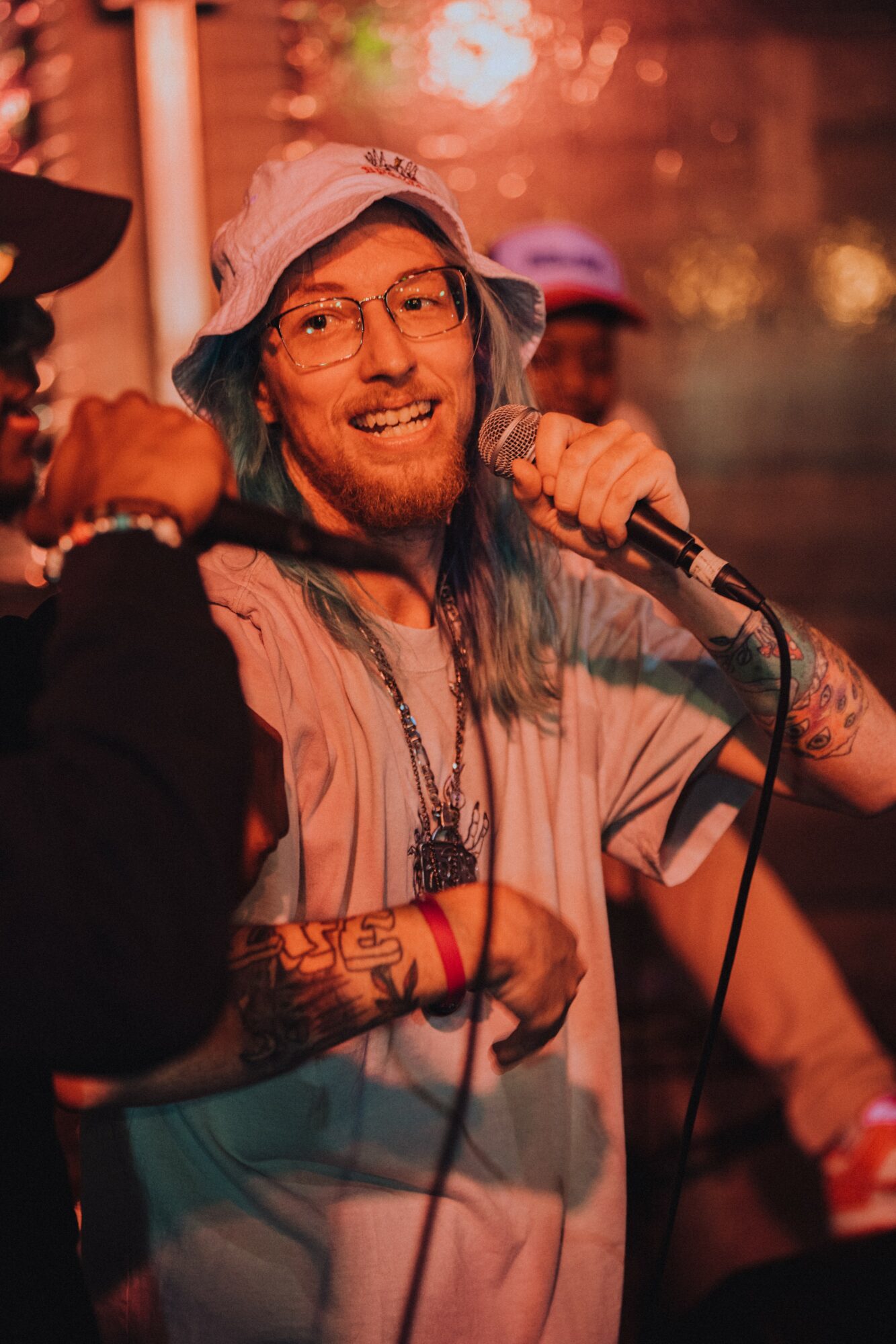 Smiling person with long hair, glasses, and tattoos holding a microphone, wearing a hat and casual clothing, in a lively setting.