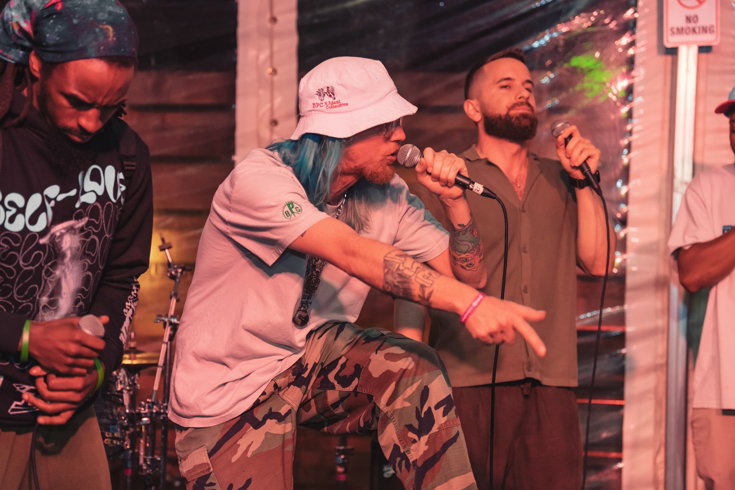 Two men singing into microphones on stage, one wearing a white hat and camouflage pants, the other with a beard, in a lively setting.