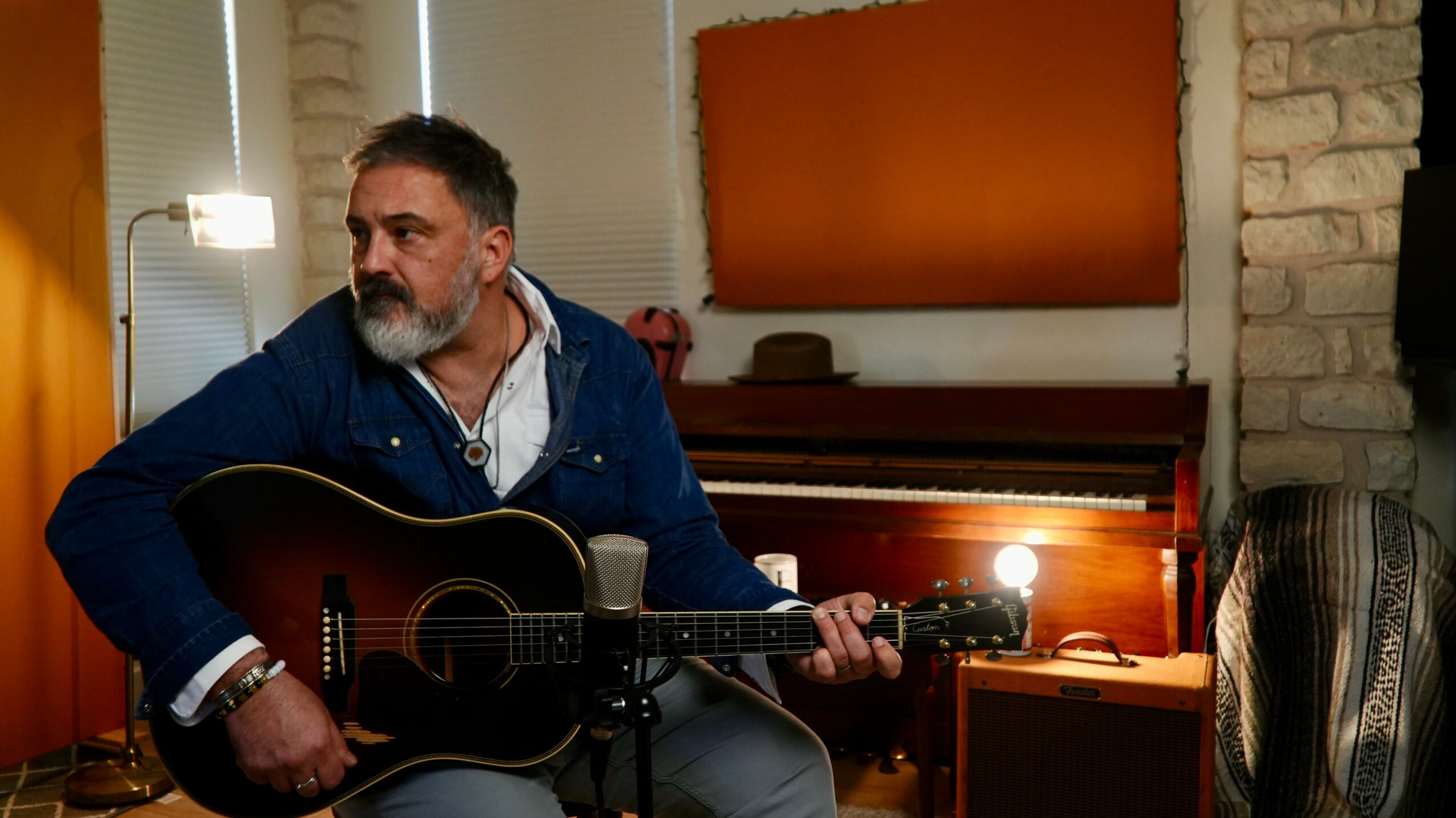 Man with a beard playing an acoustic guitar in a room with a piano and brick wall.