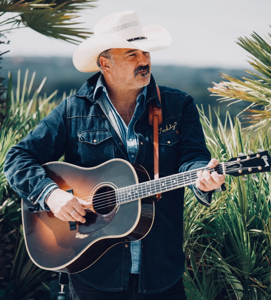 Man wearing a white cowboy hat and denim jacket playing an acoustic guitar outdoors with greenery.