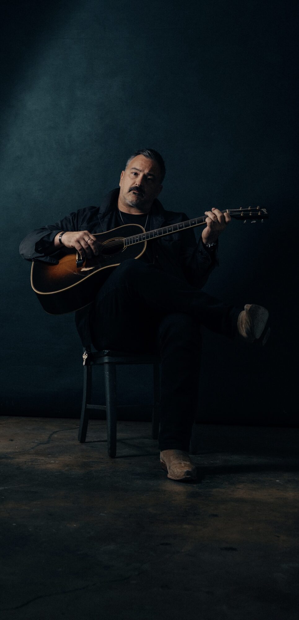 Person sitting on a chair playing an acoustic guitar against a dark background.