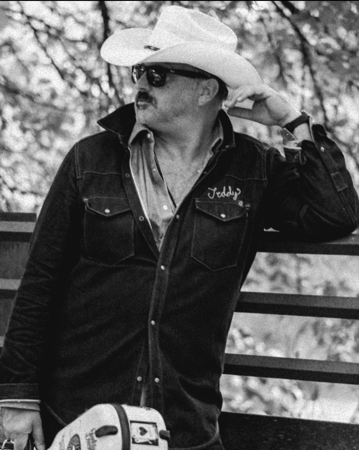 Man wearing a cowboy hat, sunglasses, and a dark jacket, leaning on a railing outdoors.