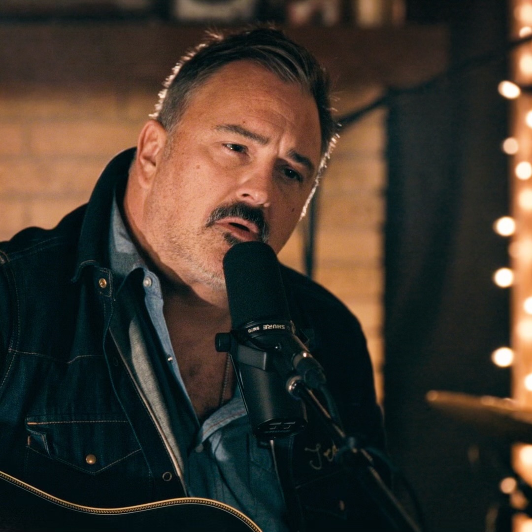 Man singing into a microphone with a serious expression, in a dimly lit setting with warm lighting.