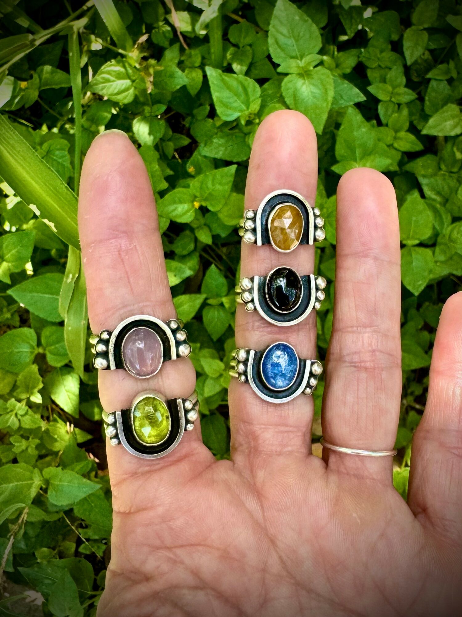 Hand with five rings, each featuring a colorful gemstone, against green leafy background.