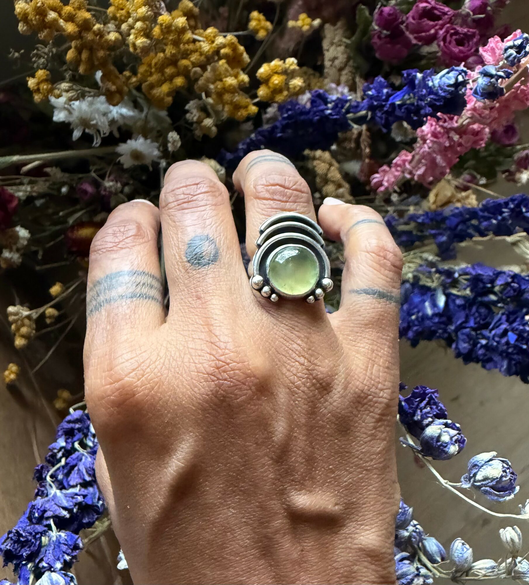 Hand with rings and tattoos, surrounded by colorful dried flowers including yellow, white, purple, pink, and blue, in a bouquet.