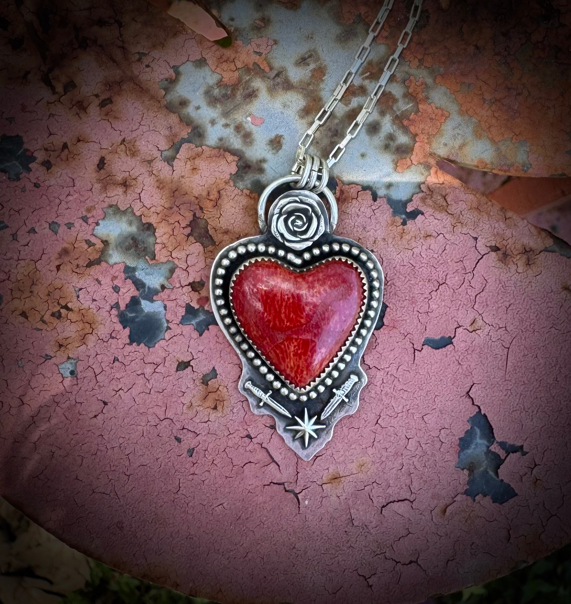Heart-shaped pendant with red center, silver border, and star design, hanging from a chain against a cracked pink background.