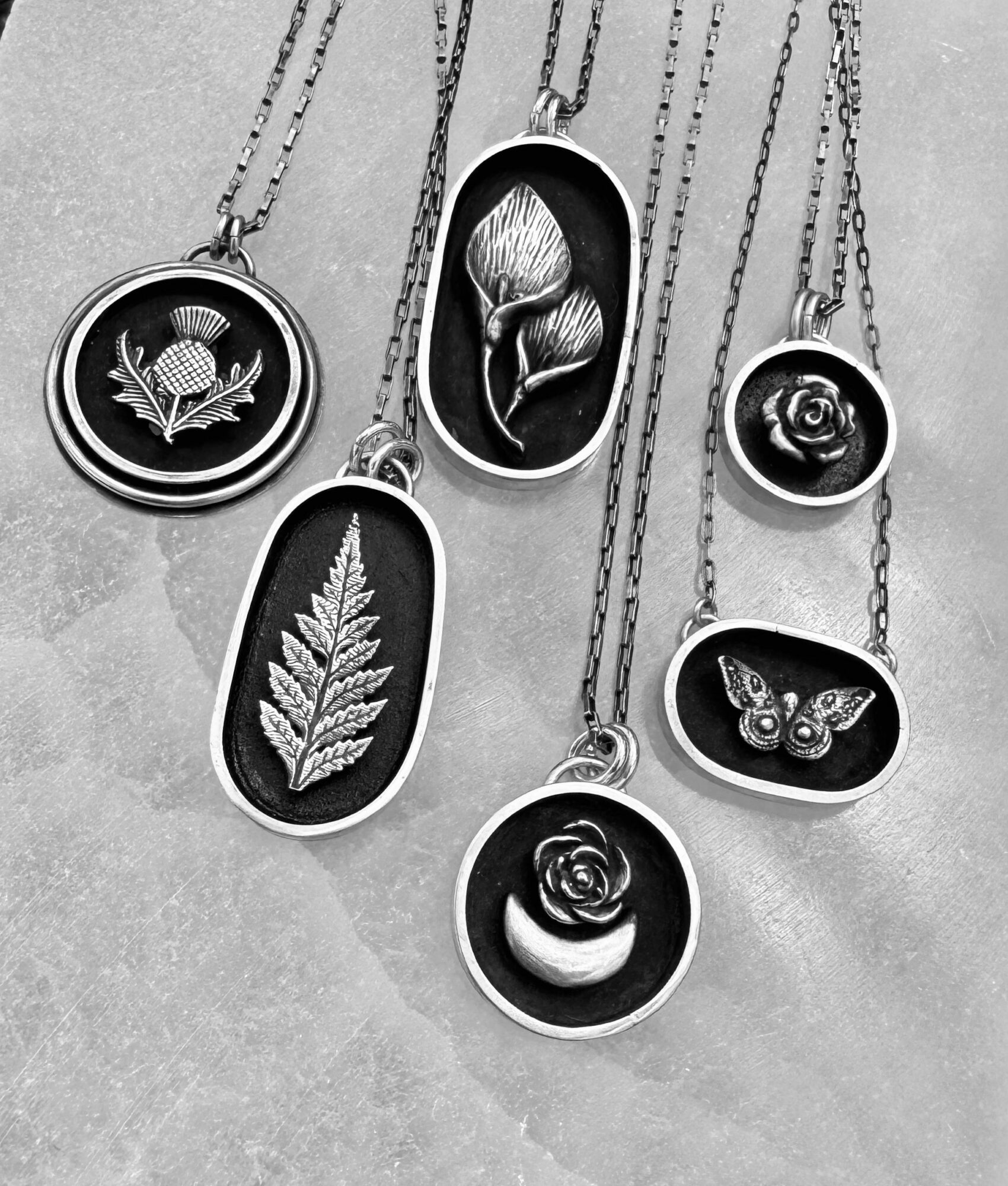 Six necklaces with various pendants, including a flower, leaf, butterfly, and rose, arranged on a textured surface.
