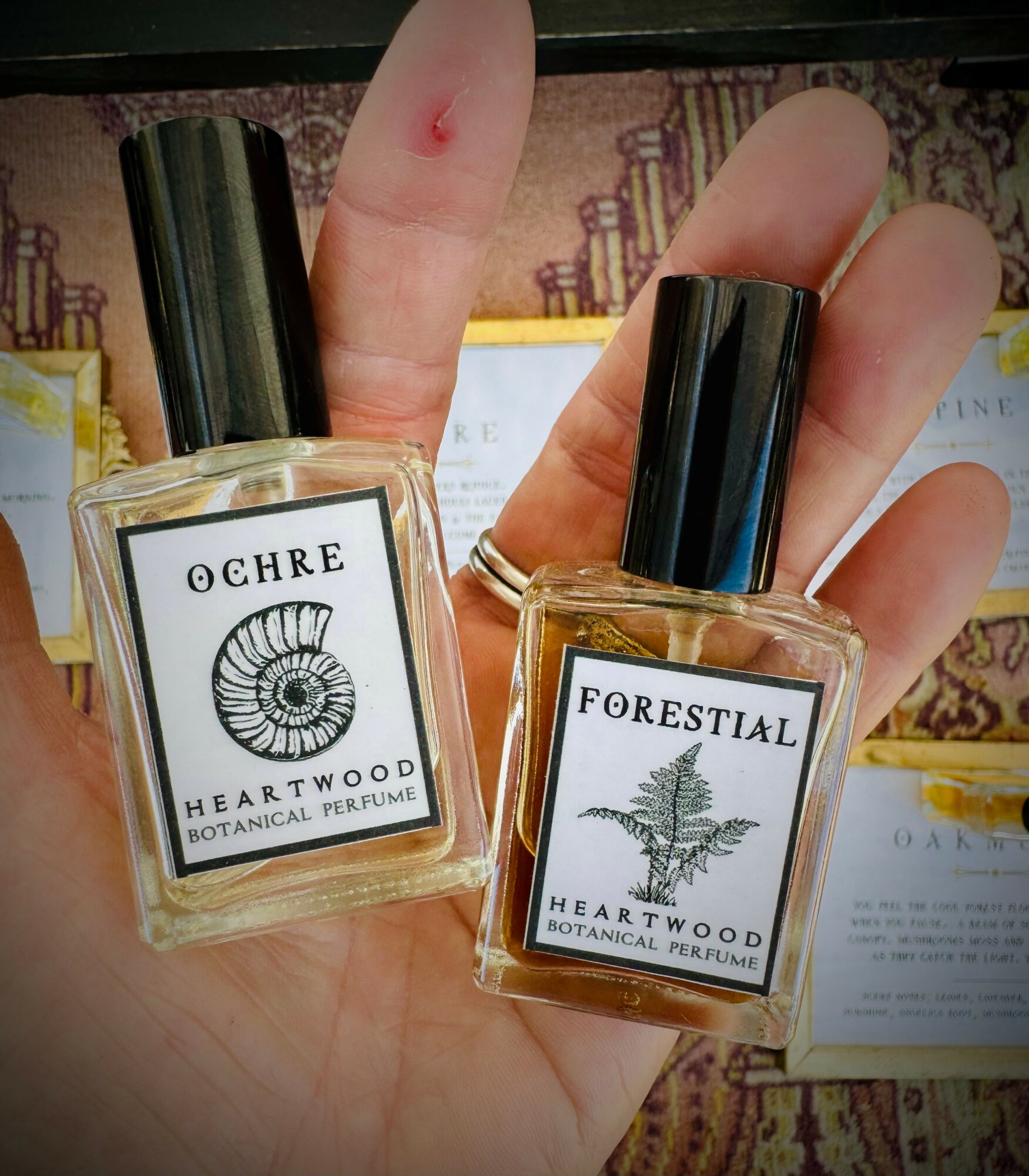 Person holding two perfume bottles labeled 'OCHRE' and 'FORESTIAL' with black caps, background shows patterned fabric and papers.