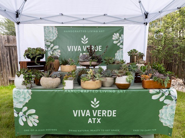 Table with potted plants under a white canopy, green banner with white text and leaf design, outdoor setting.