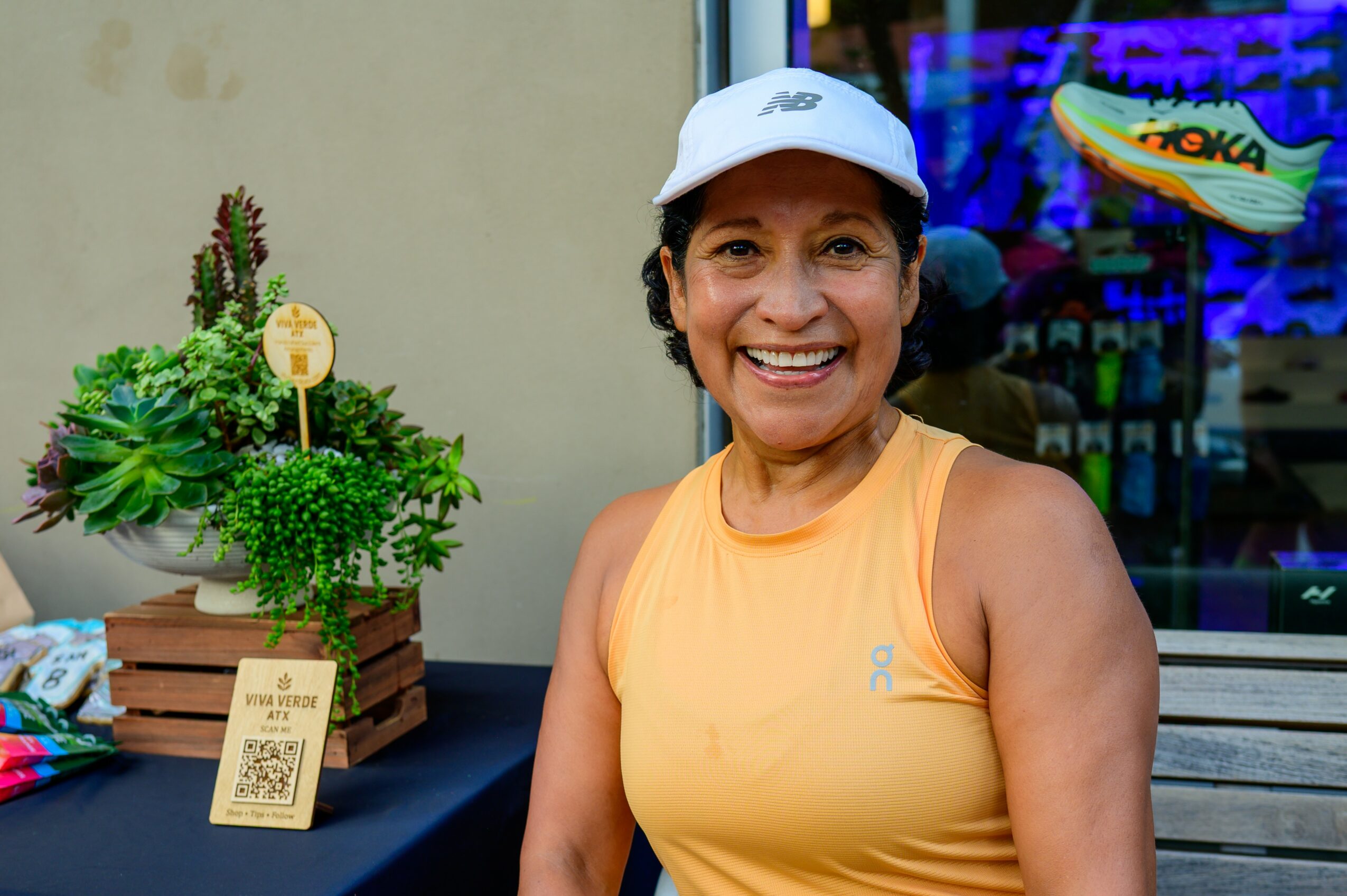 Smiling woman in yellow tank top and white cap sitting outdoors, with plants and a QR code on a table behind her.