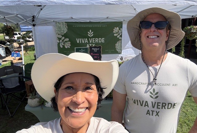 Two smiling women wearing wide-brimmed hats and sunglasses outdoors at a booth with a green sign that reads 'Viva Verde ATX'.