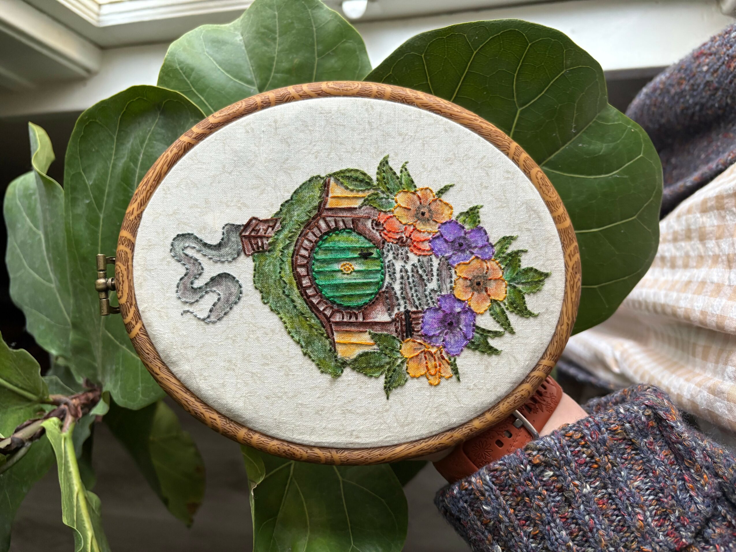 Embroidery hoop with a design of a frog sitting on a lily pad surrounded by colorful flowers, placed on green leaves.
