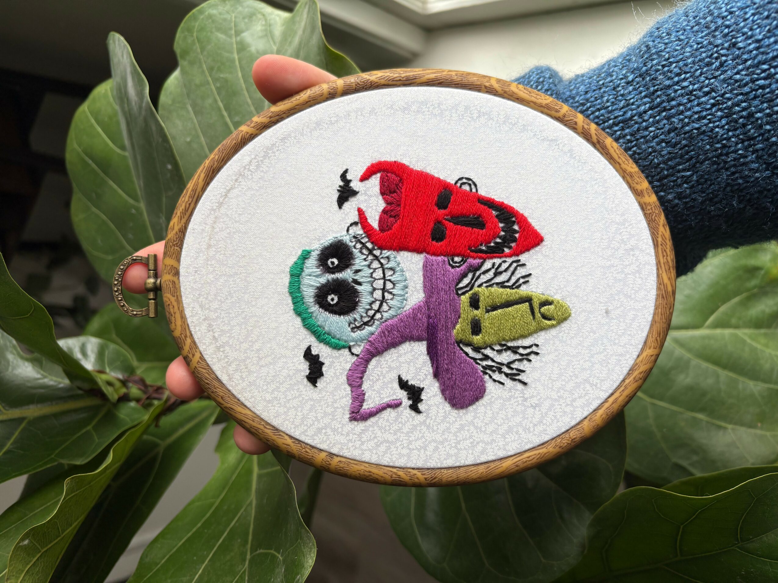 Embroidery hoop with colorful stitched monsters, held by a person wearing a blue sweater, surrounded by green leaves.