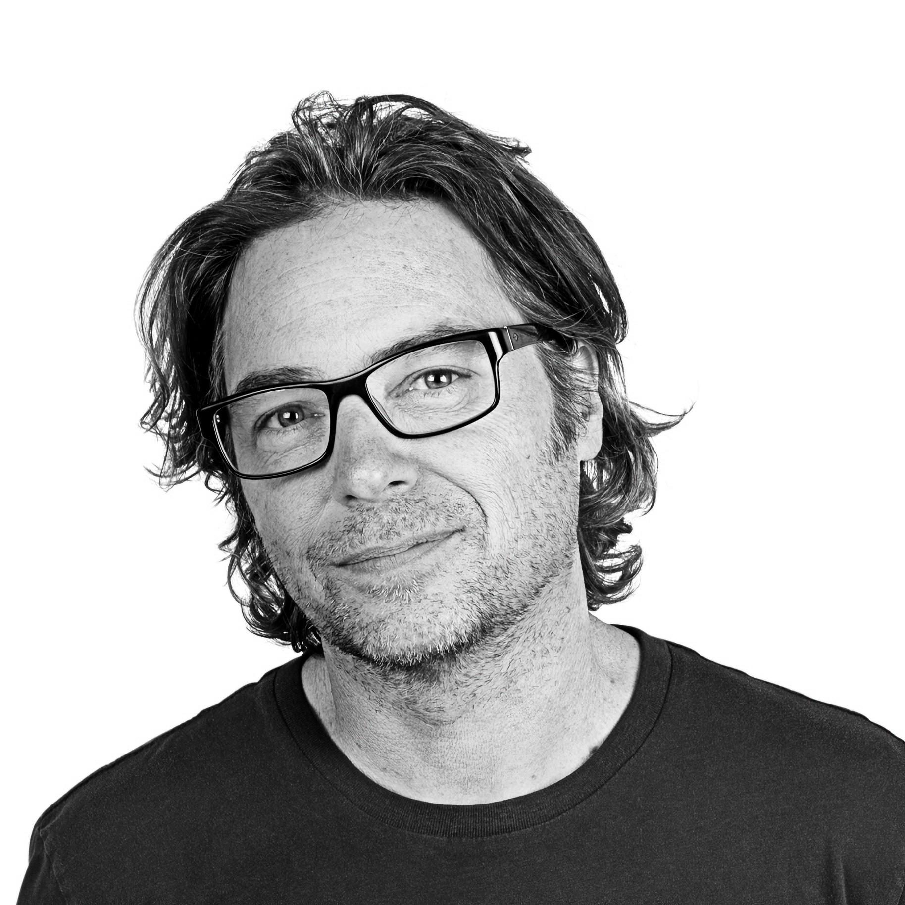 A man with glasses and medium-length hair, smiling slightly, wearing a dark shirt, against a plain background.