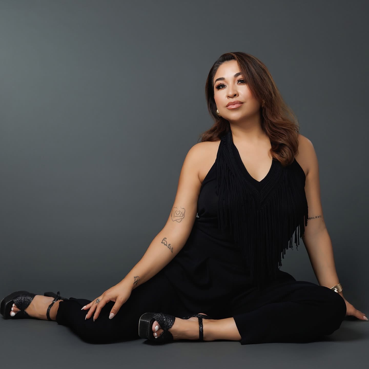 Woman sitting on the floor with legs crossed, wearing a black sleeveless top and pants, against a plain gray background.