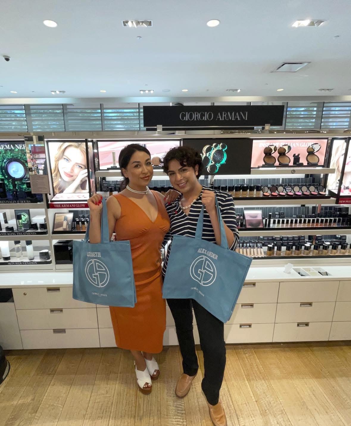 Two women stand in a cosmetics store holding shopping bags, smiling at the camera, with shelves of makeup products behind them.
