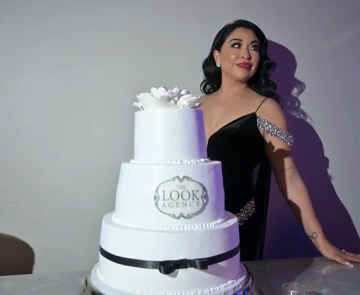 Woman in black dress standing next to a multi-tiered white cake with decorative elements, looking to the side.