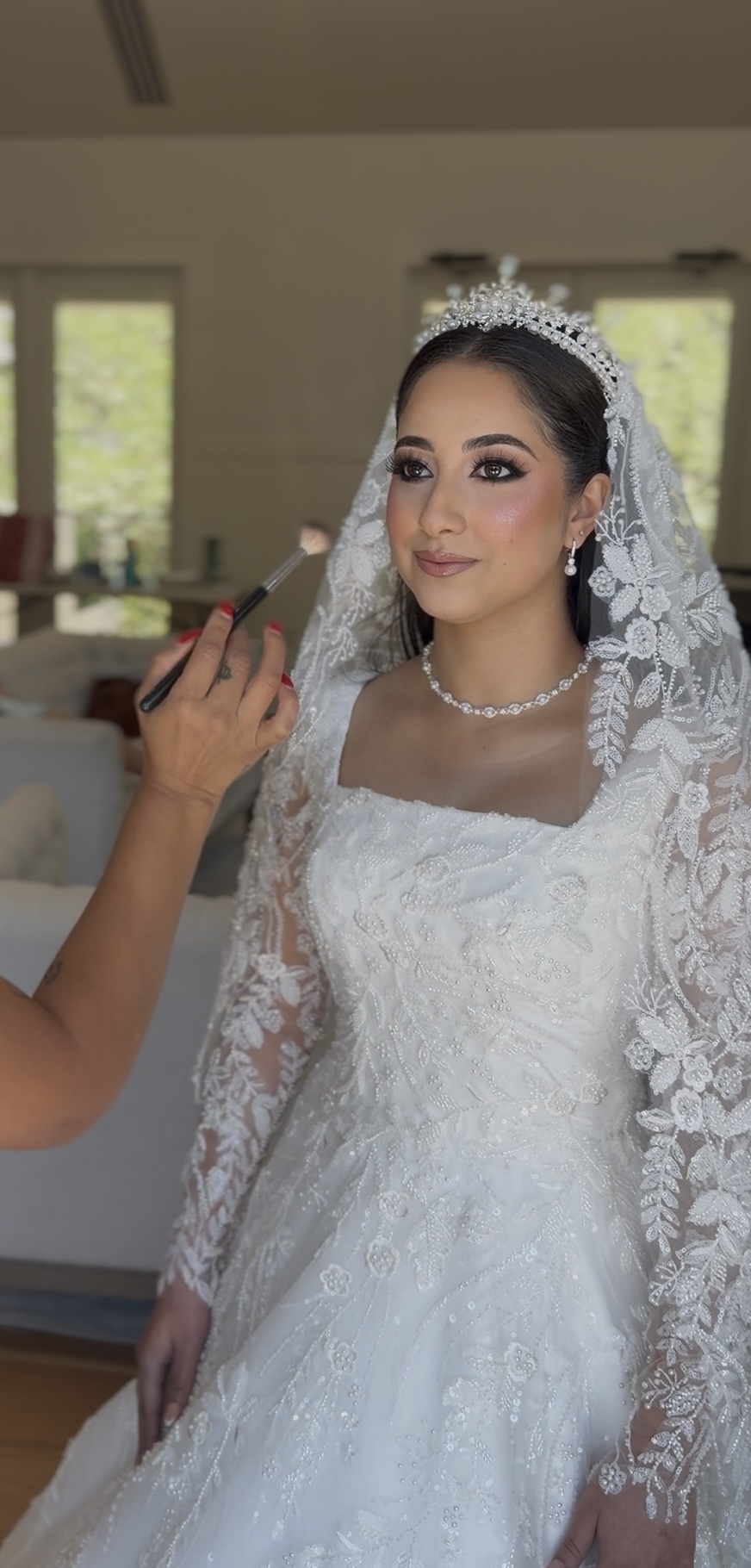 Bride in wedding dress with lace sleeves, tiara, and pearl necklace, having makeup applied, indoors with windows in background.
