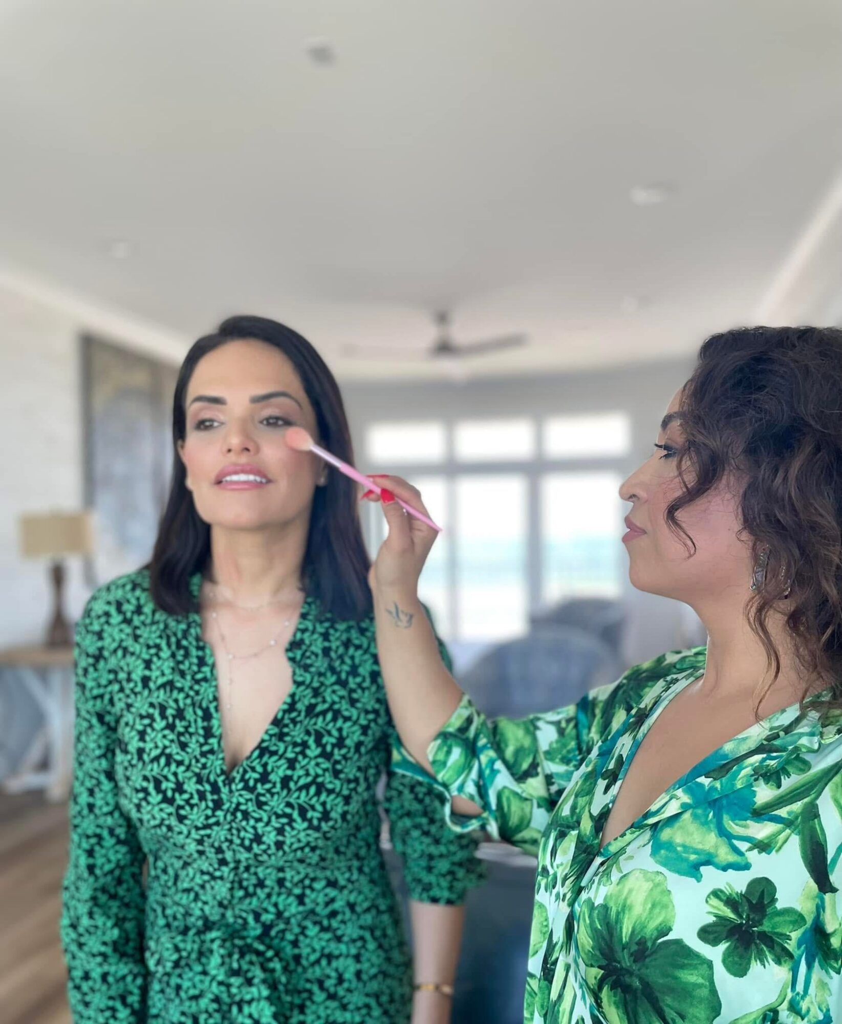 Two women in a bright room, one applying makeup to the other's face, with large windows in the background.