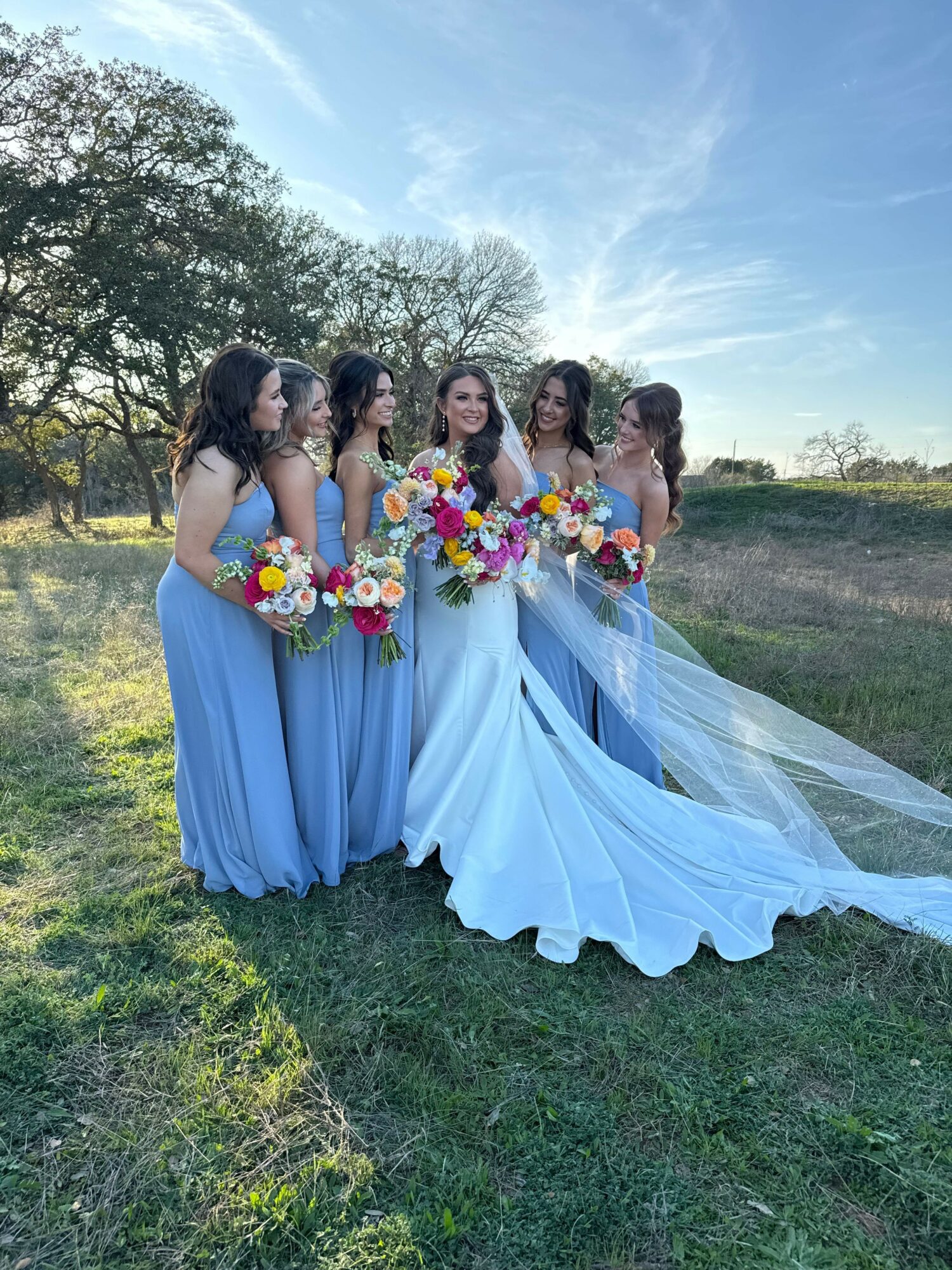 Five women in blue dresses holding colorful bouquets stand outdoors on grass with trees and sky in background.
