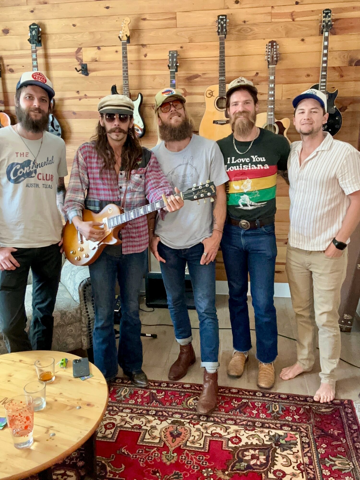 Five men standing in a room with guitars on the wall behind them, smiling, with a table and drinks in the foreground.