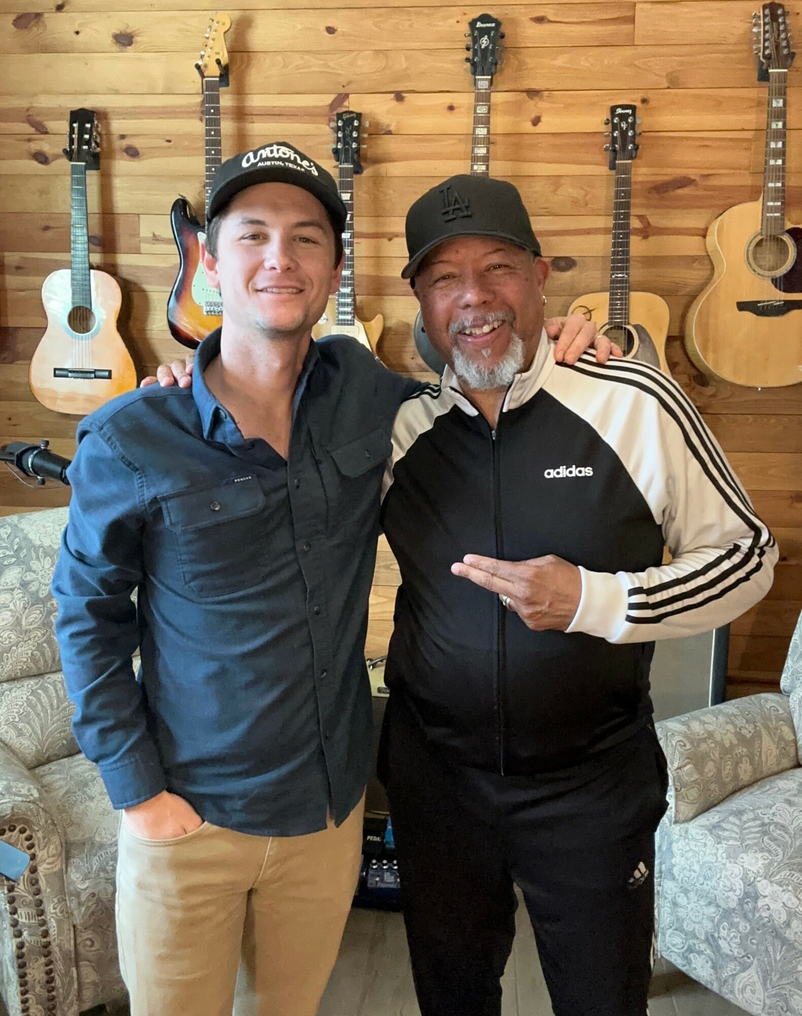 Two men smiling and posing together in front of a wall with guitars hanging, one wearing a black cap and Adidas jacket.