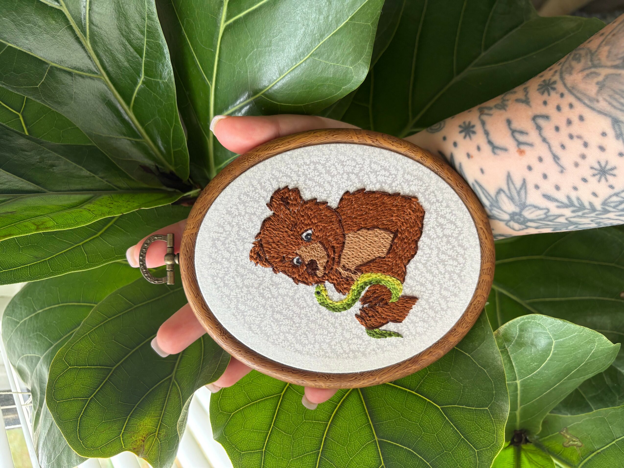 Cross-stitch embroidery of a squirrel holding a green object, on white fabric, held by a person with a tattooed arm, surrounded by large green leaves.