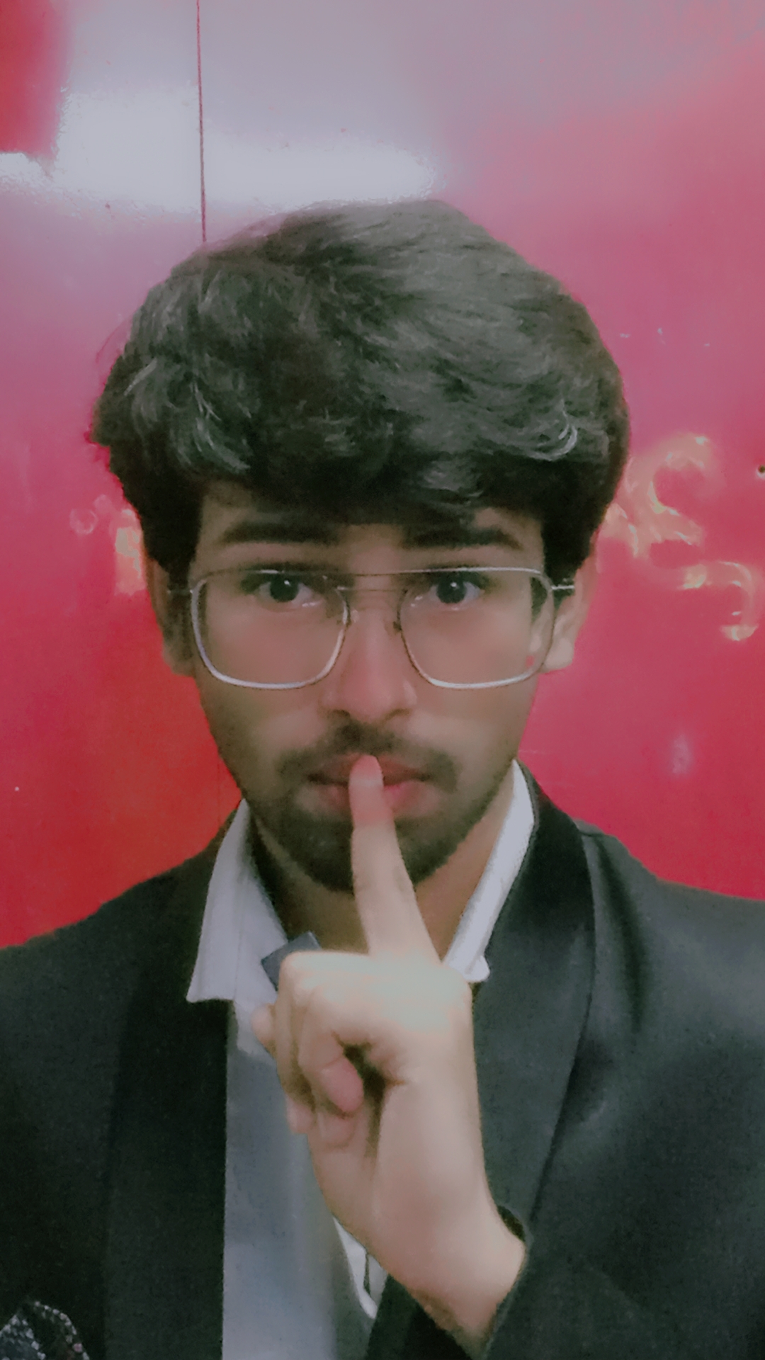Young man with glasses and dark hair holding finger to lips, wearing a suit, against a red background.
