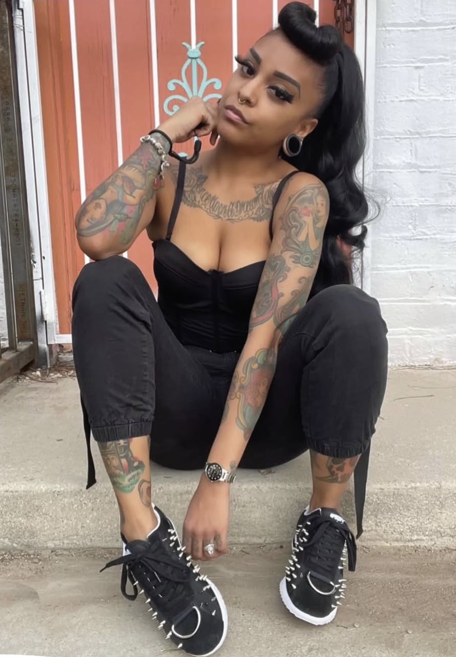 Young woman with tattoos squatting outdoors, wearing black outfit and sneakers, with a serious expression.
