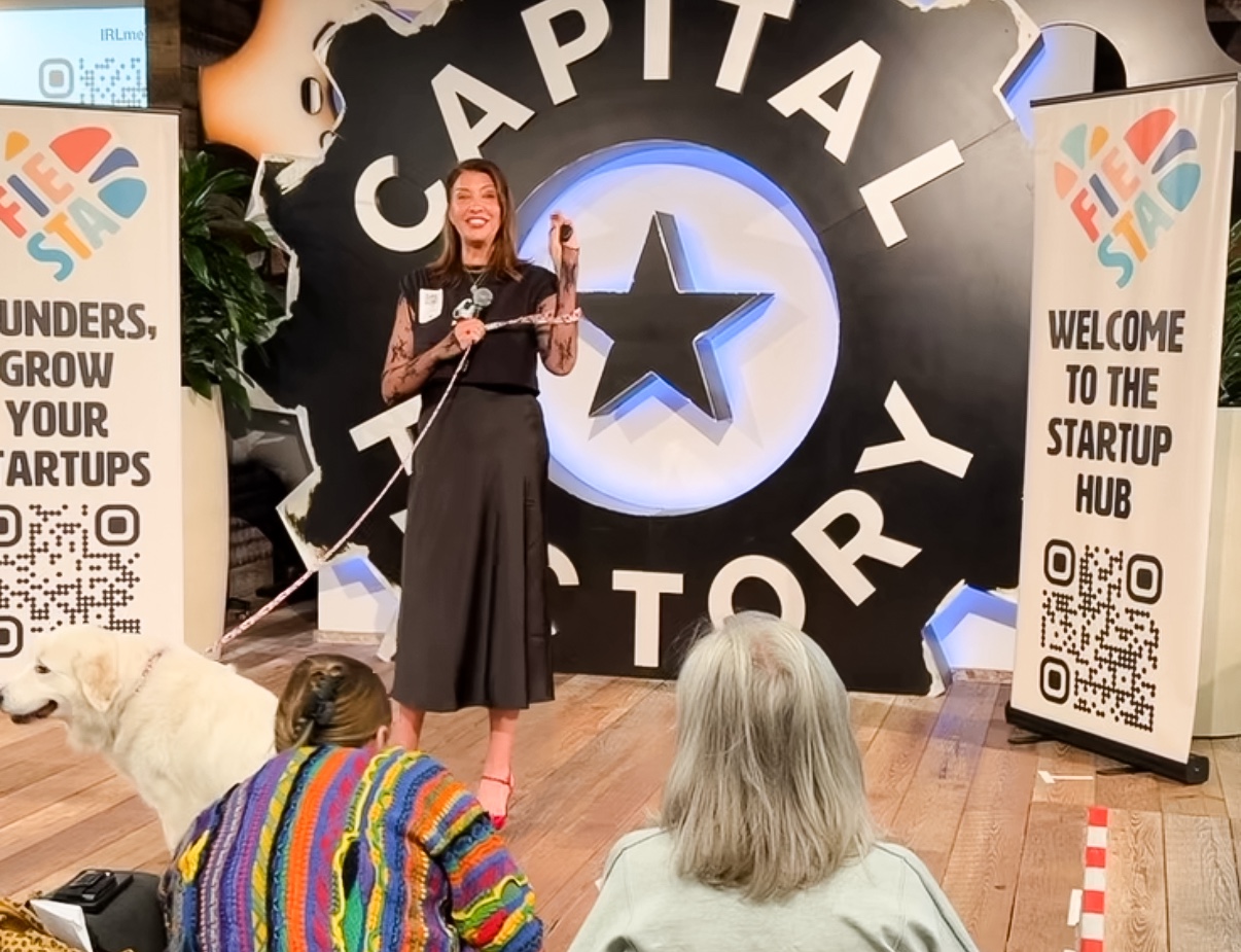 Woman speaking on stage at startup event with audience and startup hub backdrop, two vertical banners on sides, colorful sweater in foreground.