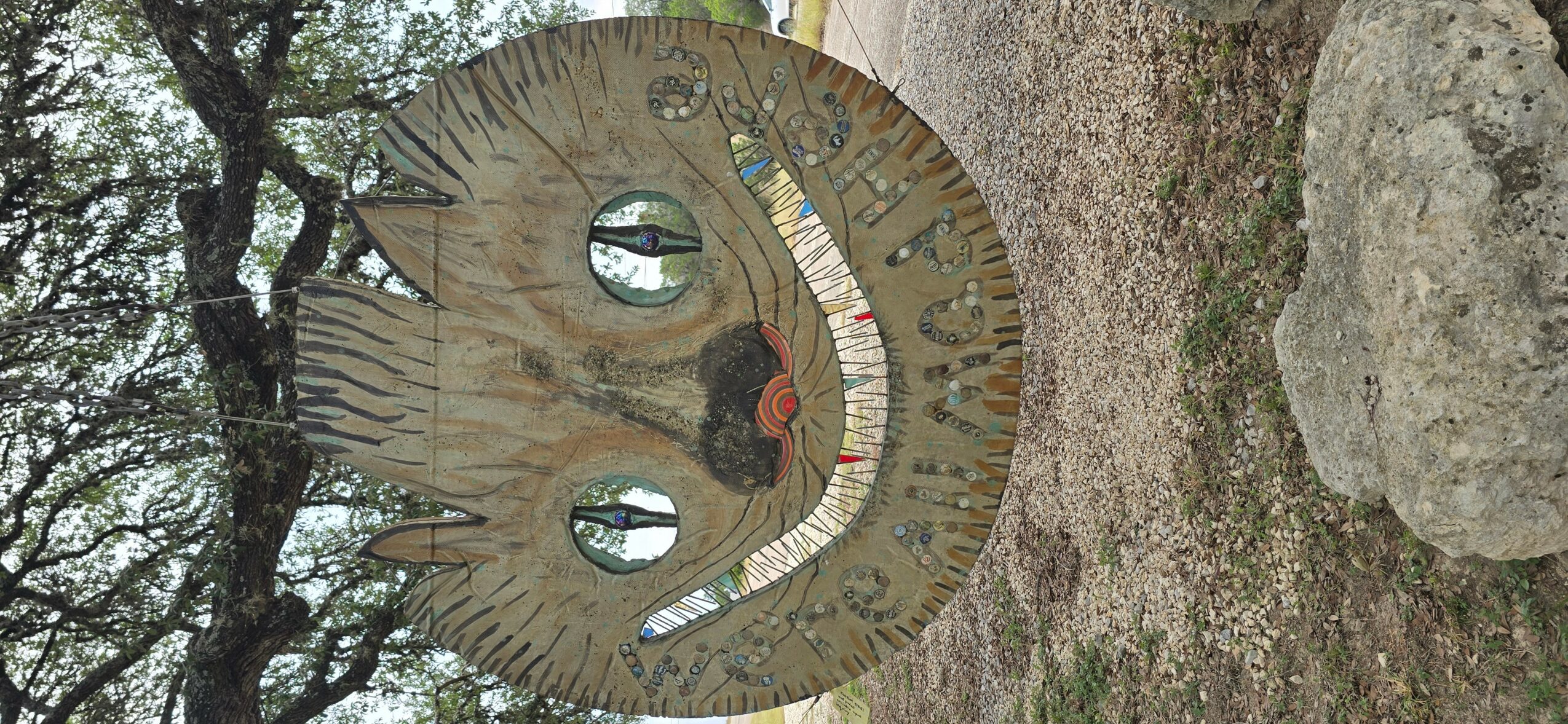Decorative circular sculpture with a smiling face, two holes for eyes, and a red tongue, outdoors with trees and rocks.