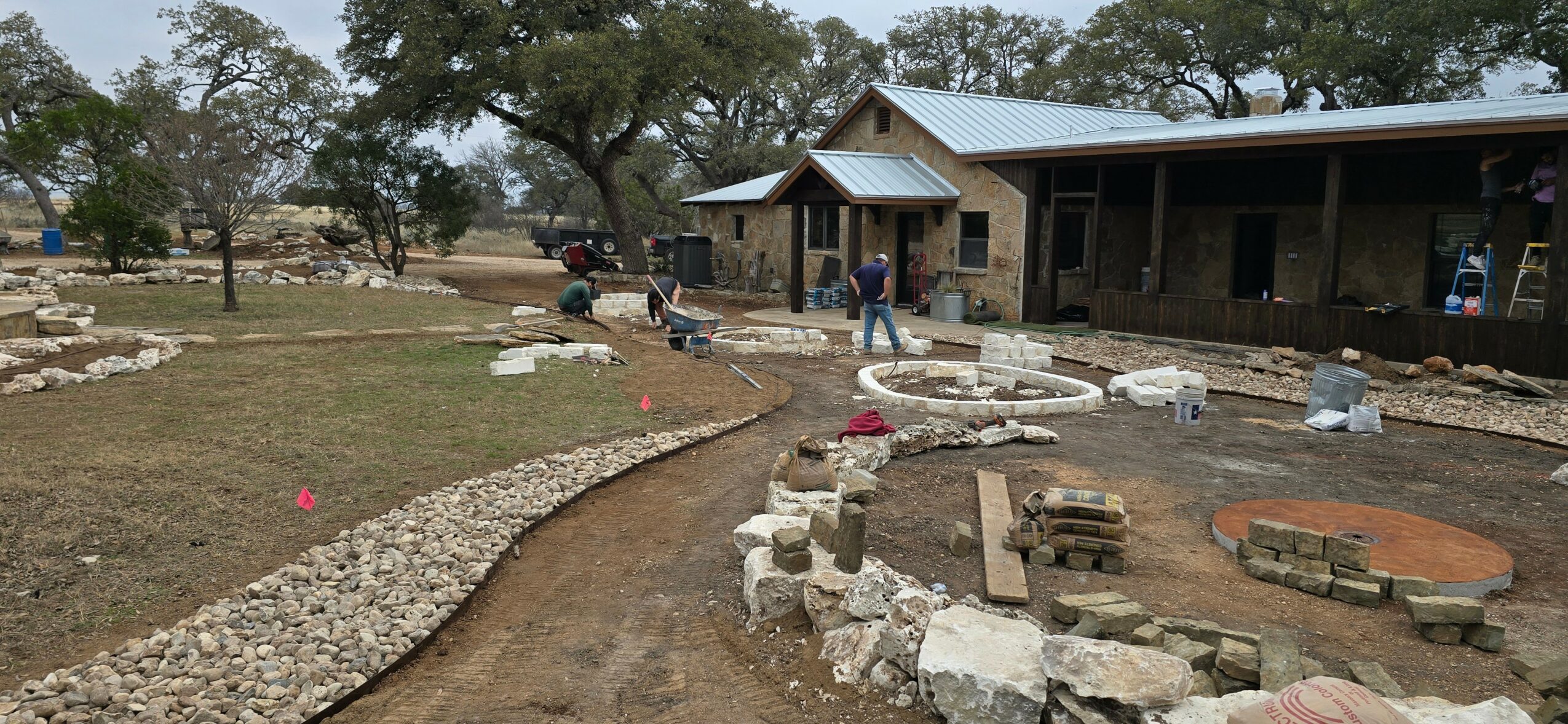 Construction site with a building, trees, and a partially completed pathway with stones and bricks.