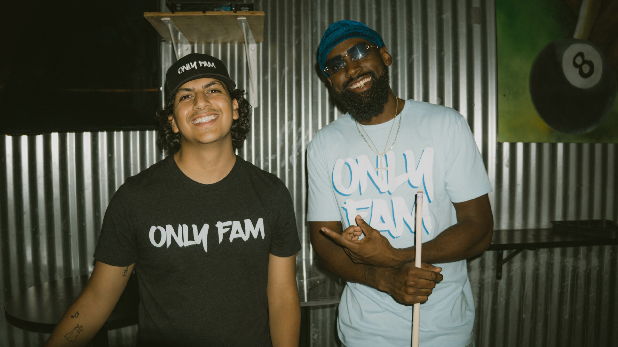 Two people smiling, one wearing a black shirt and the other a light blue shirt, in front of a metallic background.