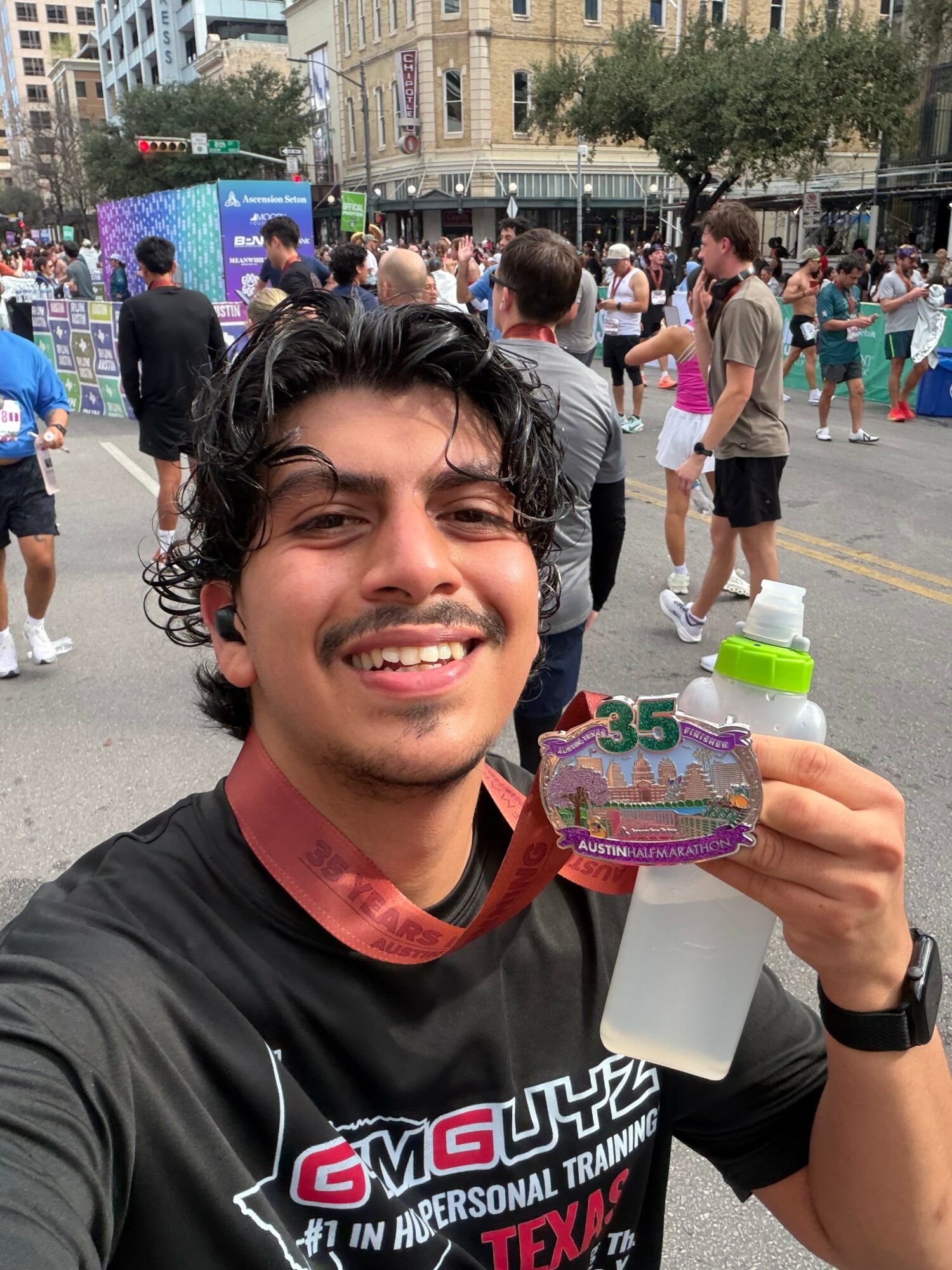 Young man smiling outdoors holding a decorated medal and spray bottle, with a crowd and city street in background.