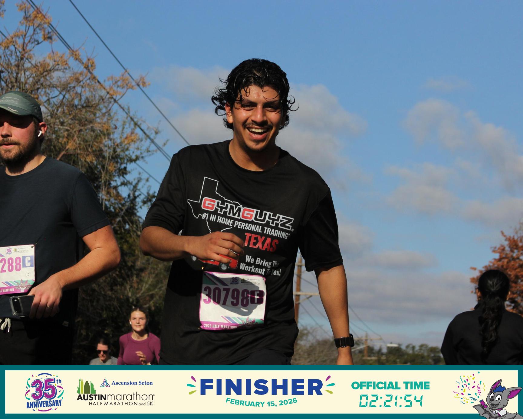 Smiling man in black shirt holding a race bib, running outdoors with blue sky and trees in background.