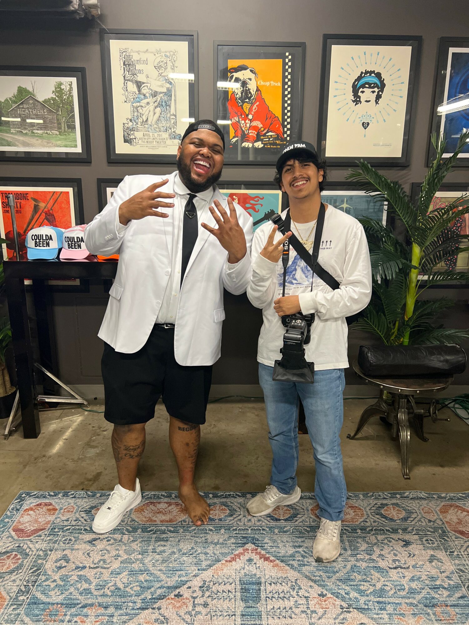 Two people smiling and making peace signs in front of framed artwork, one wearing a white jacket and shorts, the other in a white jacket and jeans.