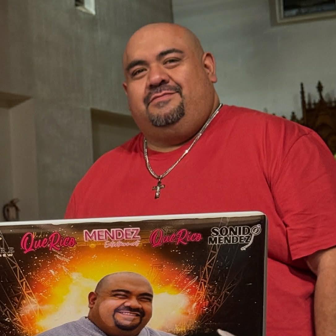 Man with a beard and mustache wearing a red shirt and a cross necklace, holding a laptop with a bright background, smiling.