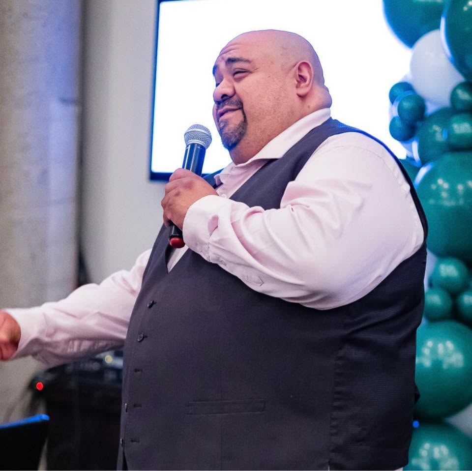 Man holding microphone, speaking at event, with balloons and screen in background.
